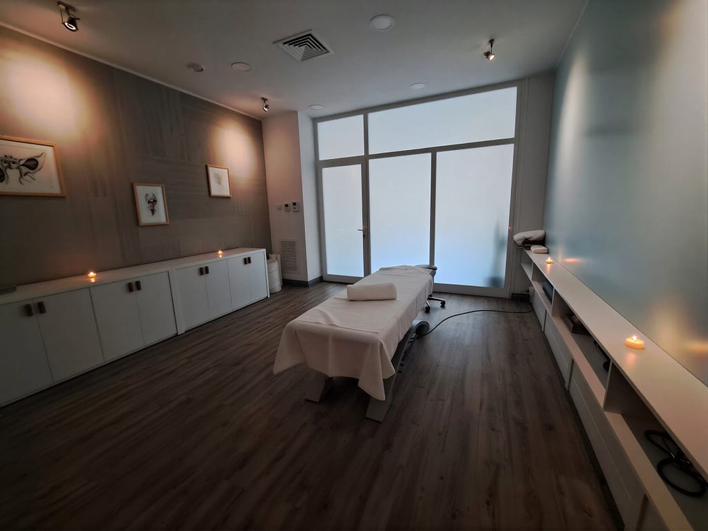 This a picture go a practice room, soften light ans candles on the shelves. The bench is prepared for a massage with towels on top
There are some big windows in the background
The blue wall brings a calming atmosphere