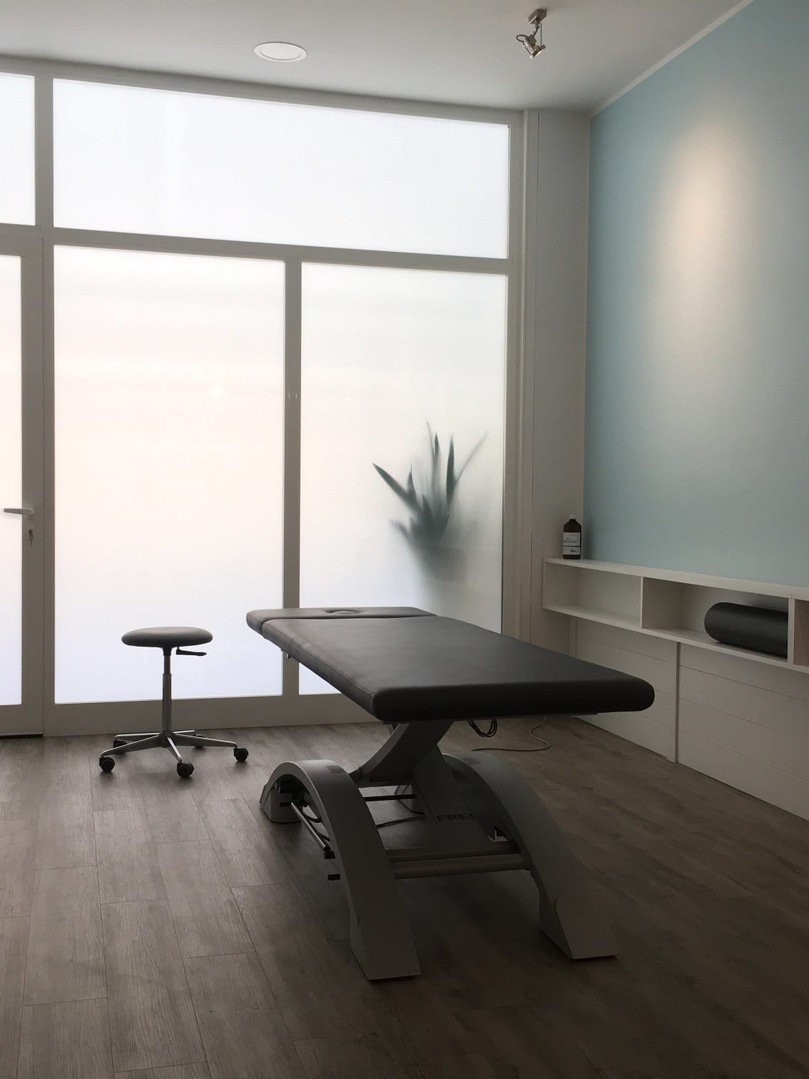 Picture of the inside of the treatment room. You can see the bench and the stool and the big windows behind. The blue wall brings a calming atmosphere