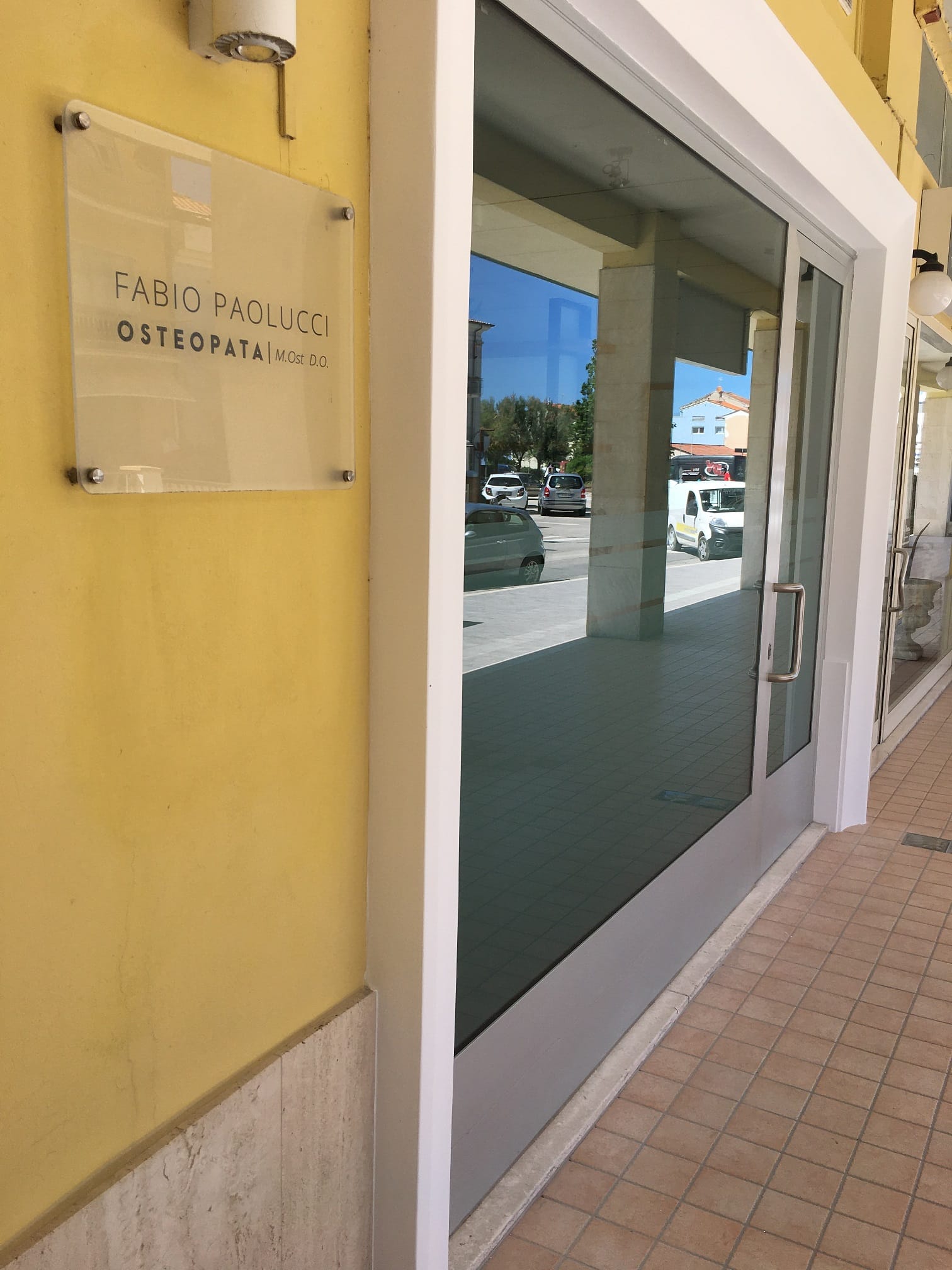 Picture taken from the left hand side of the clinic. You can see the plaque the written the name Fabio Paolucci. You can see the window and the entrance door of the practice. The walls are yellow. The picture is taken from the street