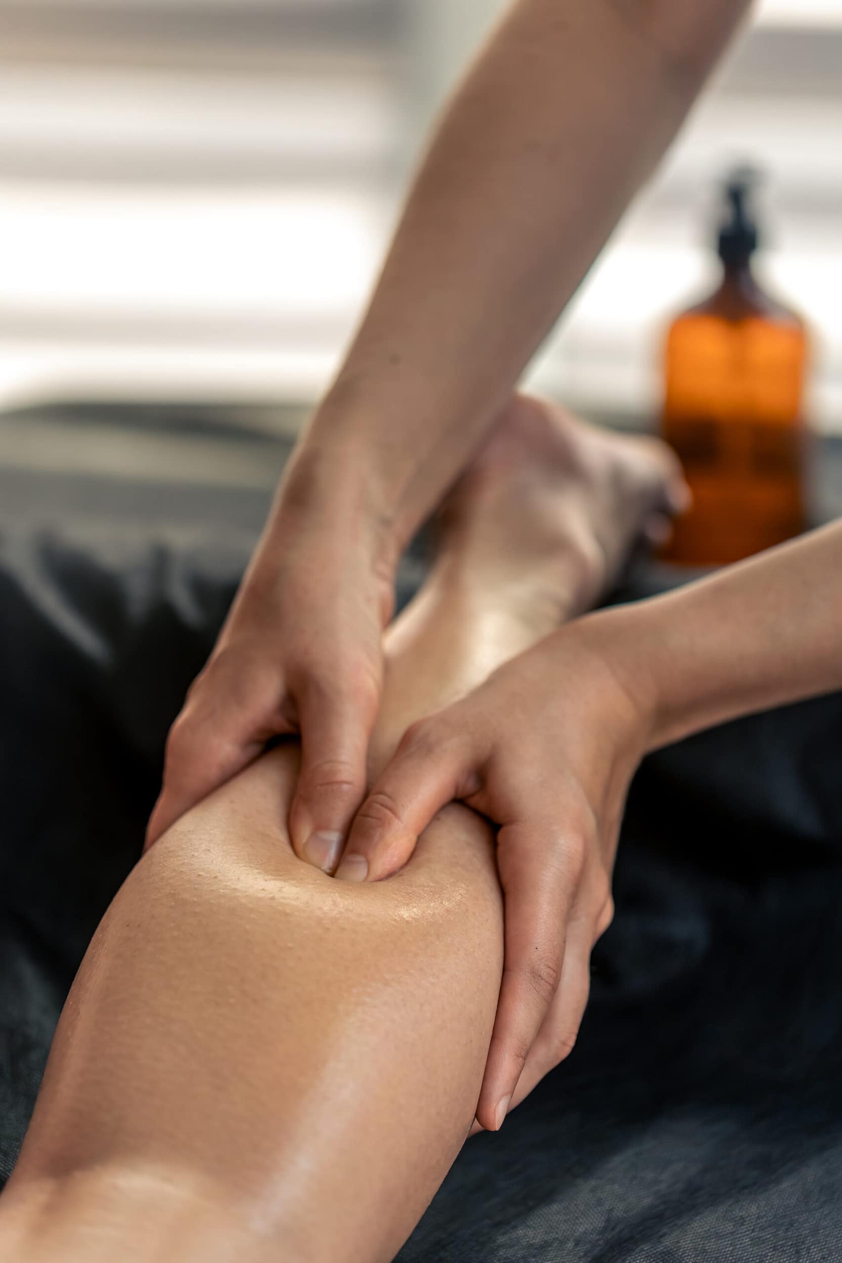 Picture of a massage made of the calf using thumbs. The atmosphere is calm and relaxing. You can also see the bottle of oil in the background